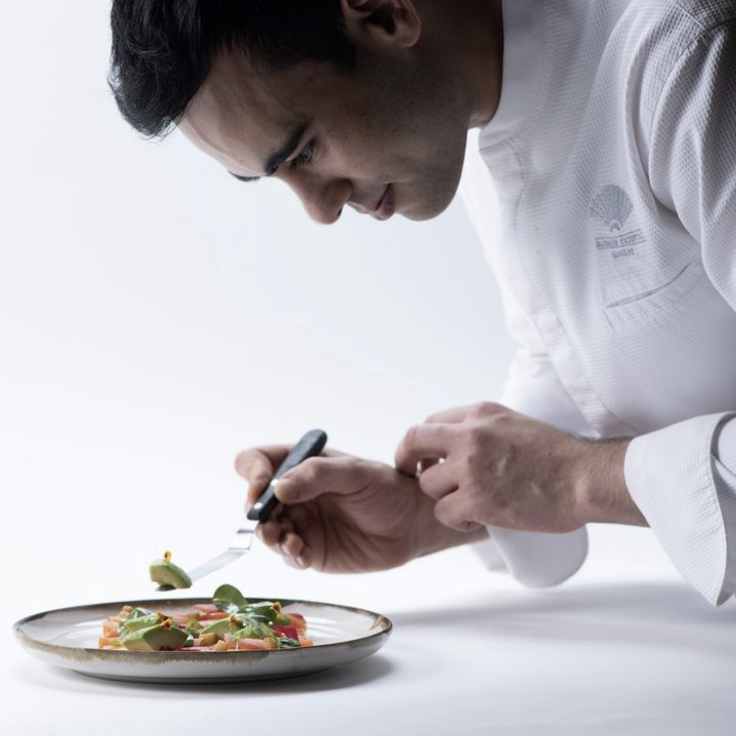 A man in a white chef's coat is taking a photo of a plate of gourmet food with a smartphone. The plate contains a colorful dish with various ingredients, garnished with herbs and vegetables.