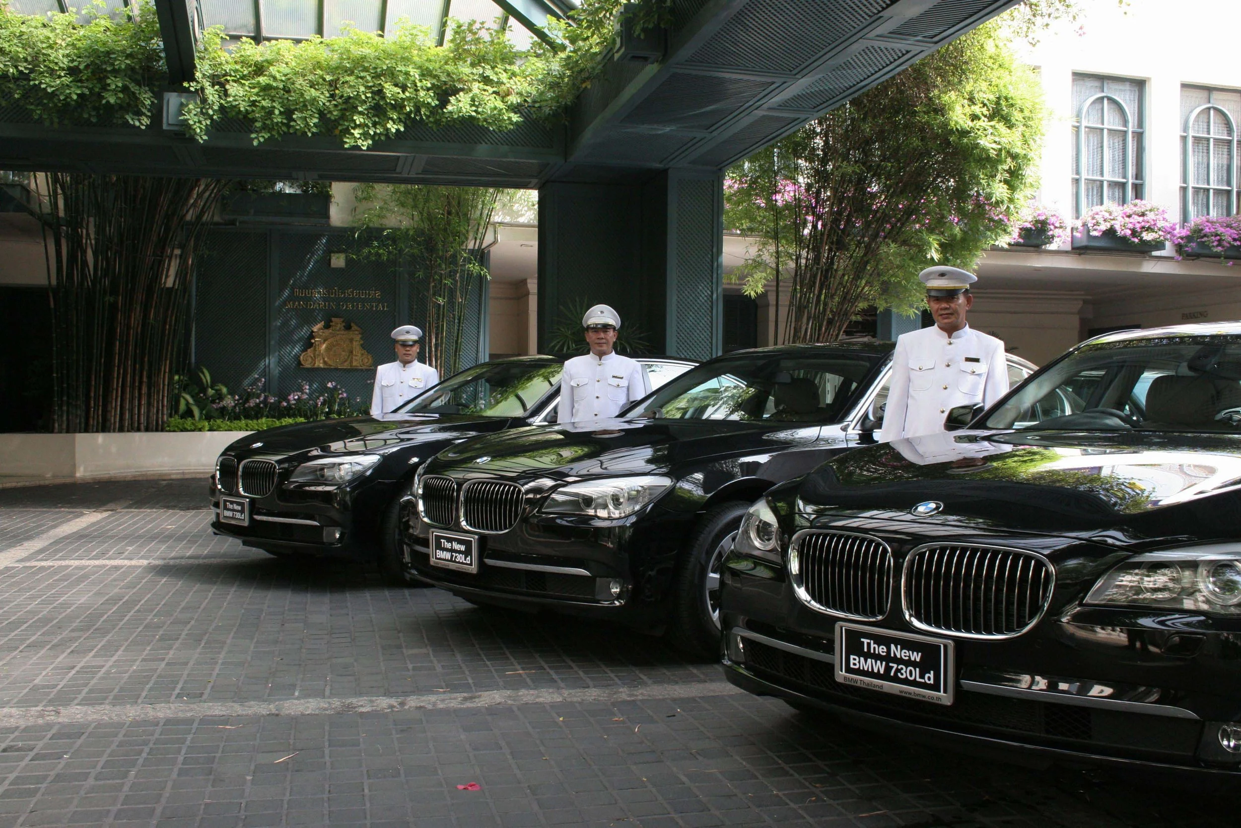 Three black BMW 730Ld cars parked in front of a building, with three uniformed men standing beside each car, two wearing white caps and one without a cap. The setting appears to be outside a hotel or similar establishment, with greenery and a sign in
