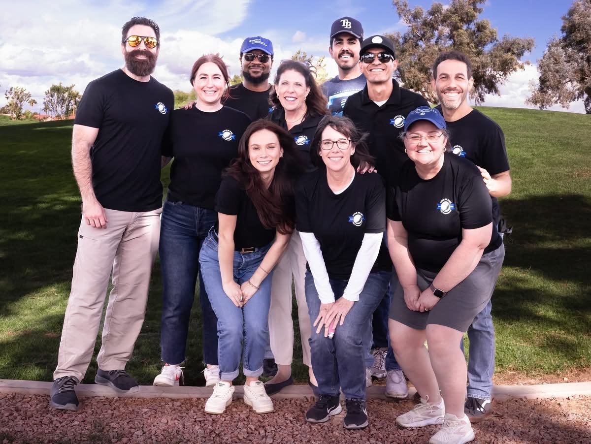 A group of ten smiling people wearing matching black shirts with a space-themed logo, standing outdoors on a grassy area with trees and a cloudy sky in the background.