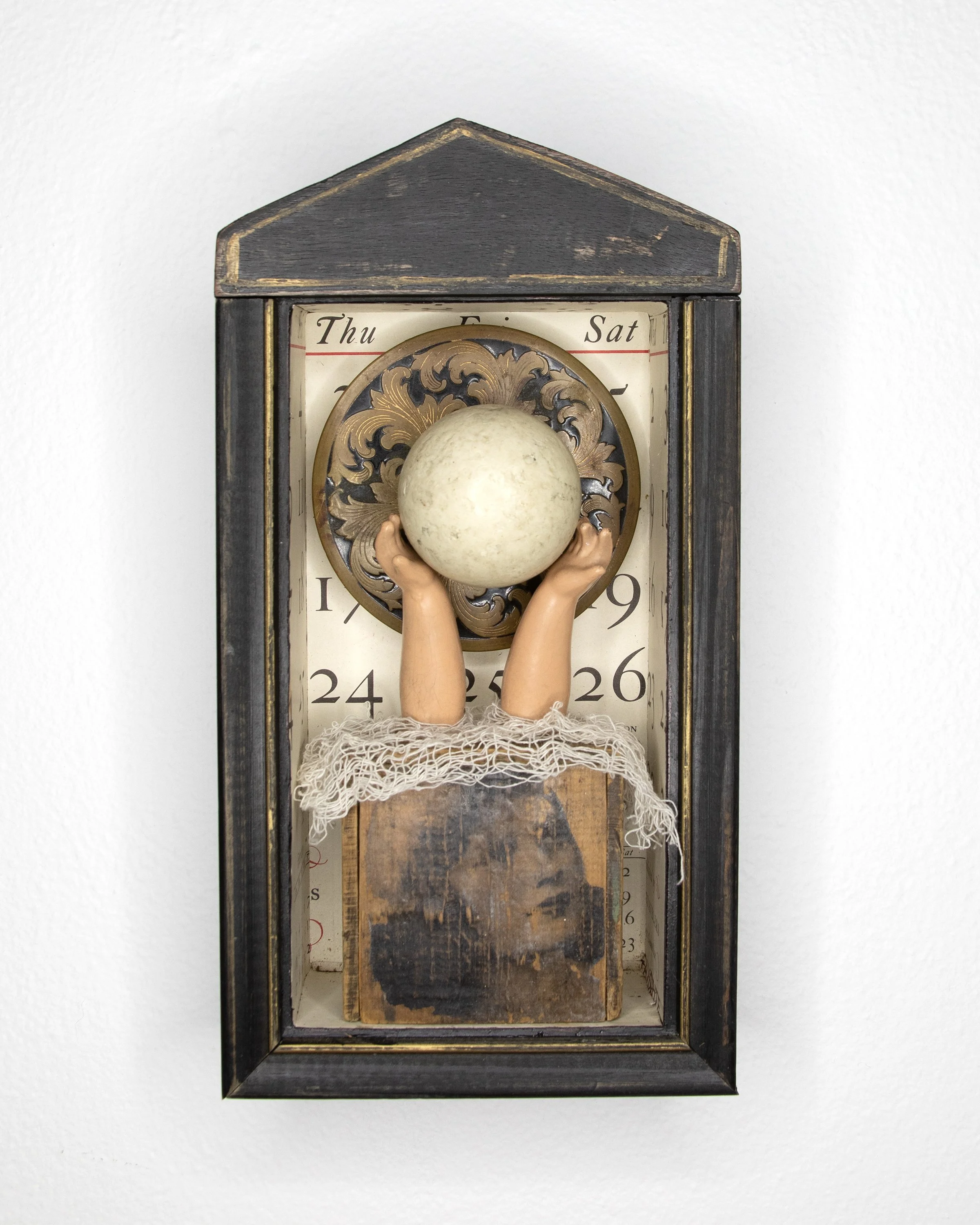 Assemblage art box featuring a vintage calendar background, a woman's portrait, doll arms lifting a white moon, and decorative antique elements within a black shadow box frame.