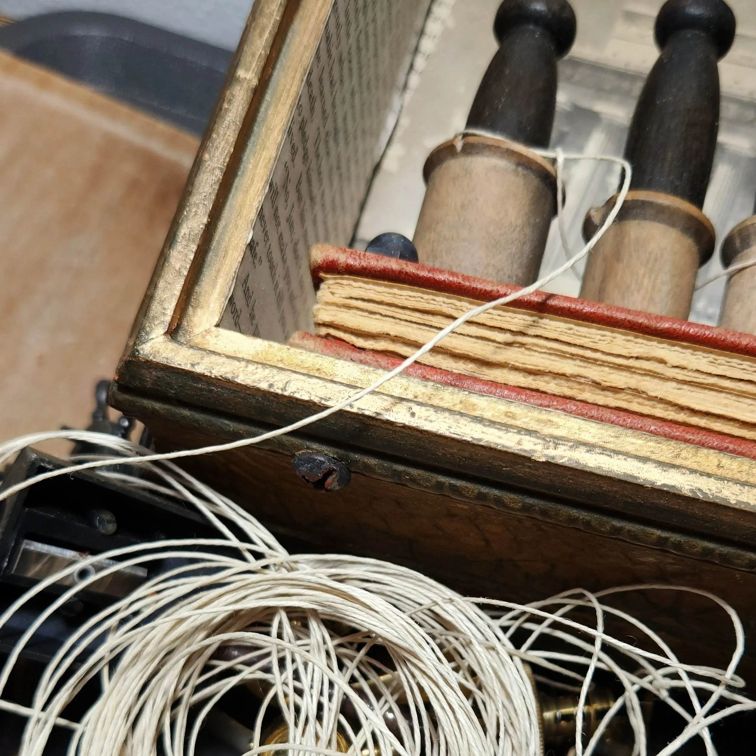 Close-up of an artist’s work area with a coil of pale cord tangled among vintage doll parts, metal hardware, thread spools, and a small assemblage box lined with printed paper.