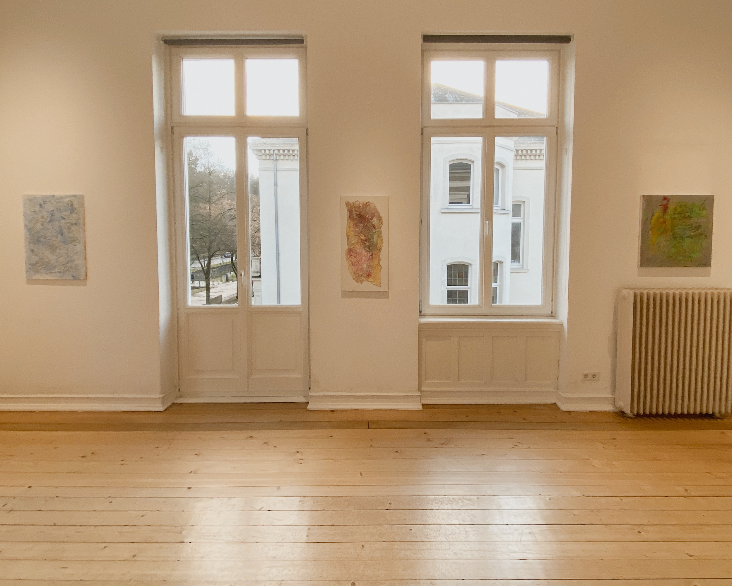 Ein leeres Zimmer mit Holzboden, weißen Wänden, drei große Fenster und einige Bilder an der Wand.