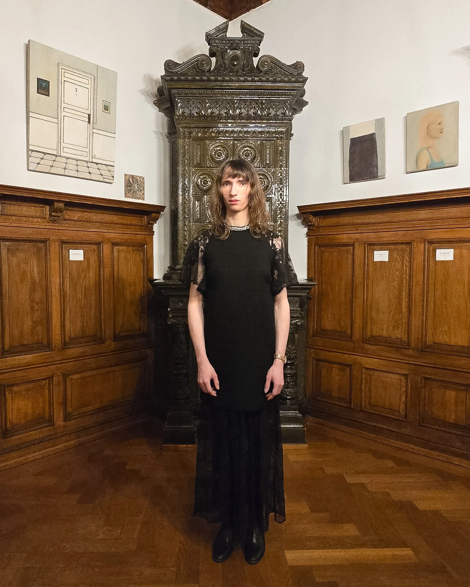 Eine junge Frau in einem schwarzen Kleid mit Spitzenärmel steht vor einer antiken, verzierten schwarzen Holzofen im Raum mit Holzverkleidung und Kunstdrucken an den Wänden.