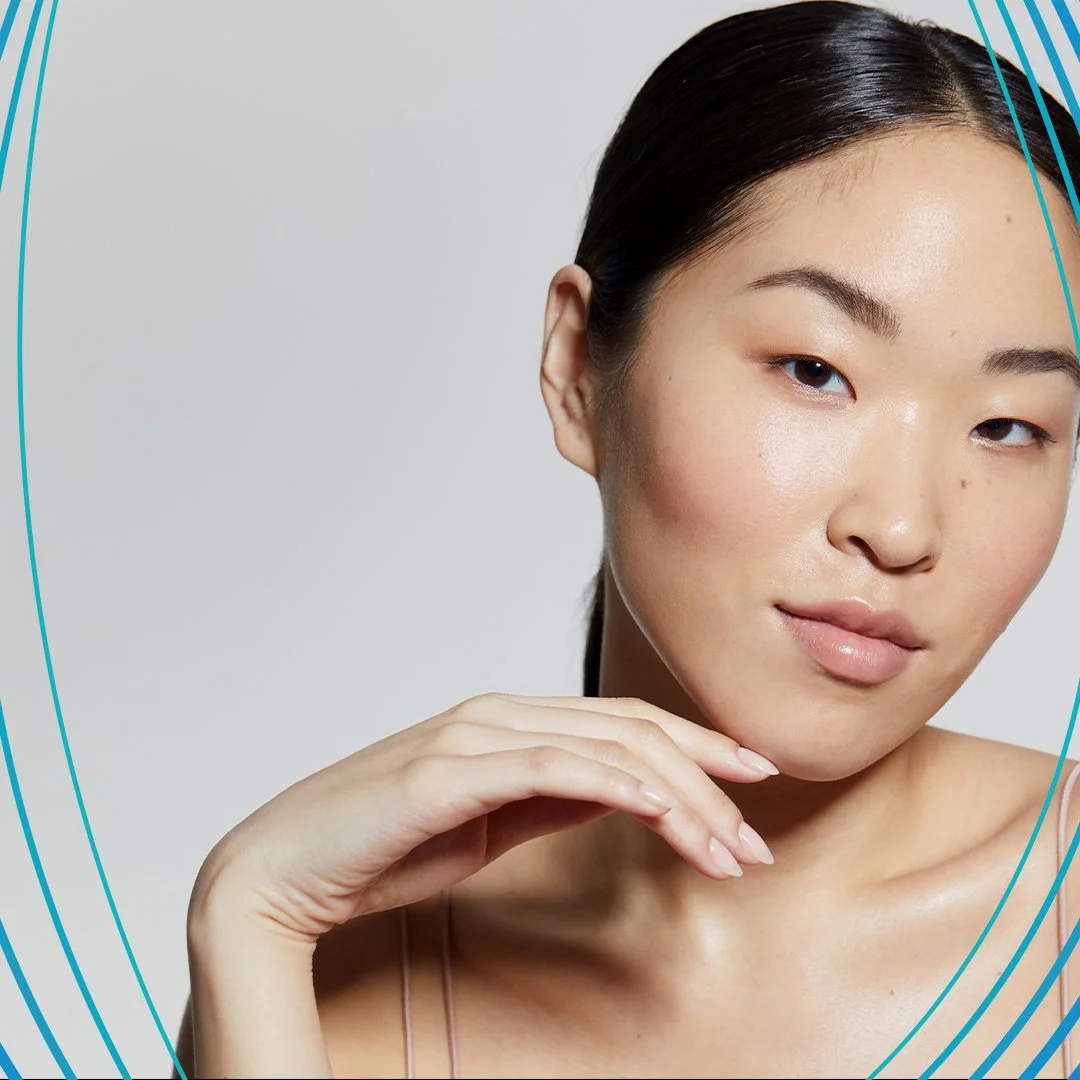Close-up of a young Asian woman with flawless skin, dark hair slicked back, and a subtle smile, posing against a plain light gray background.