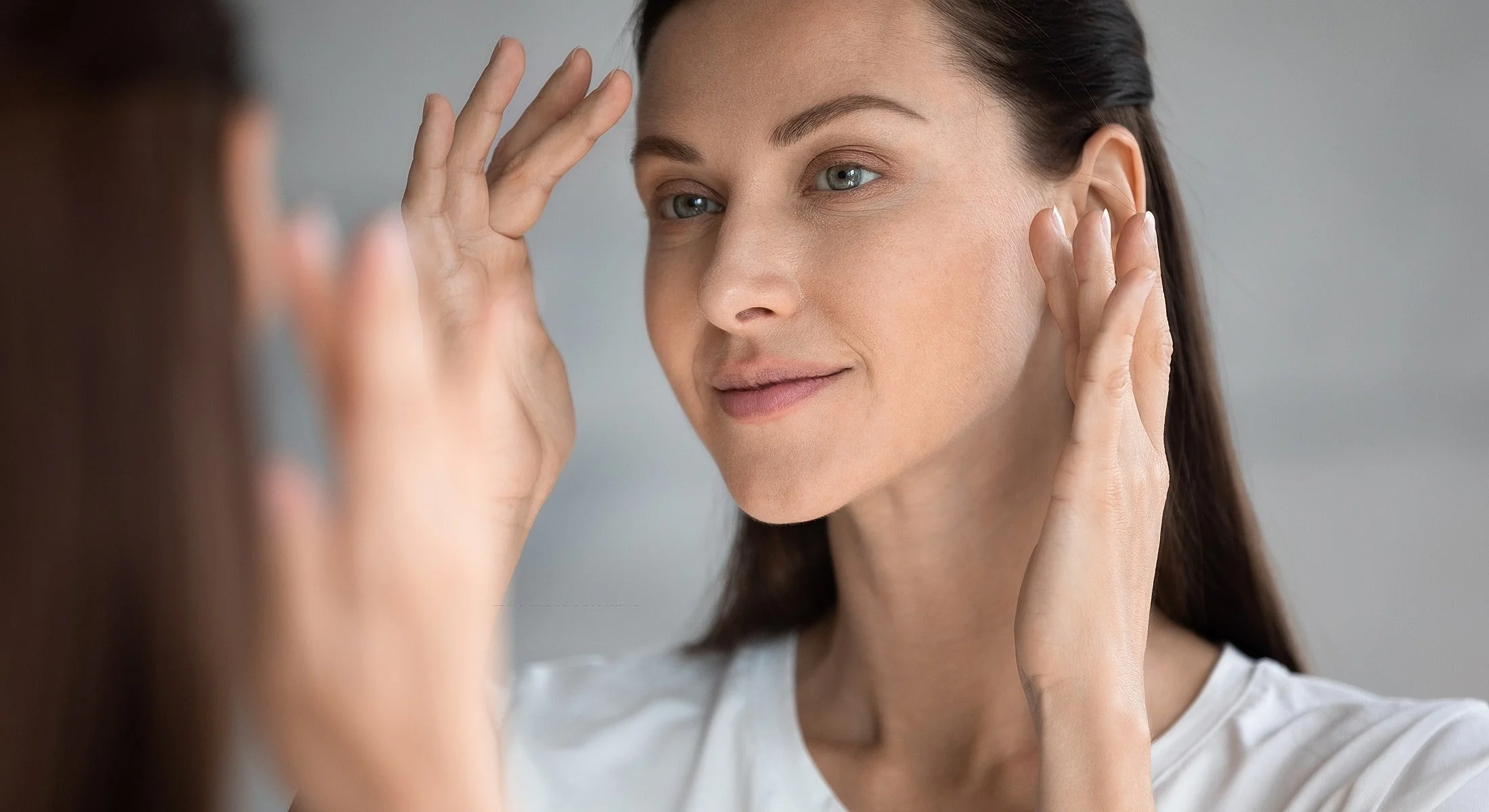 A woman gently touching her face, looking at her reflection in the mirror with a subtle smile.