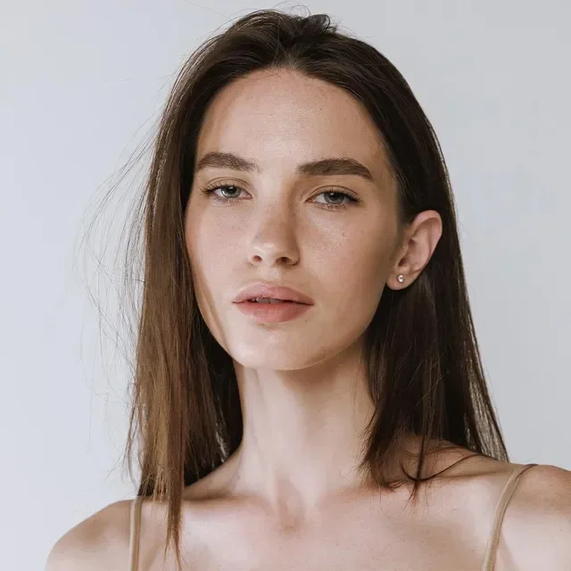 Close-up of a woman with long brown hair, fair skin, and light-colored eyes, wearing a nude-colored spaghetti strap top, with a neutral expression against a light background.