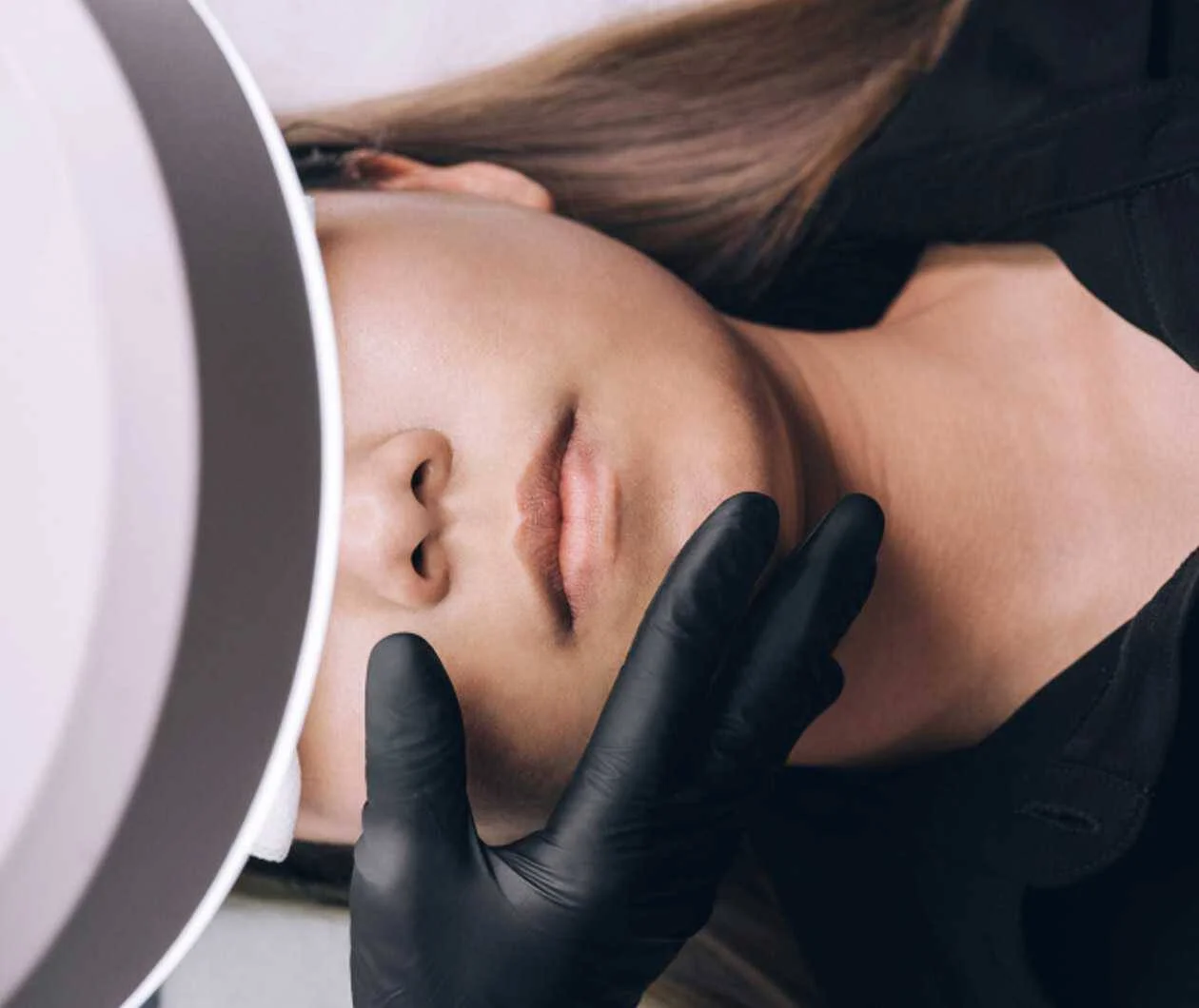 A woman receiving a cosmetic or dermatological facial treatment with her eyes closed, lying down with her head under a magnifying lamp, and a gloved hands gently touching her chin or jawline.