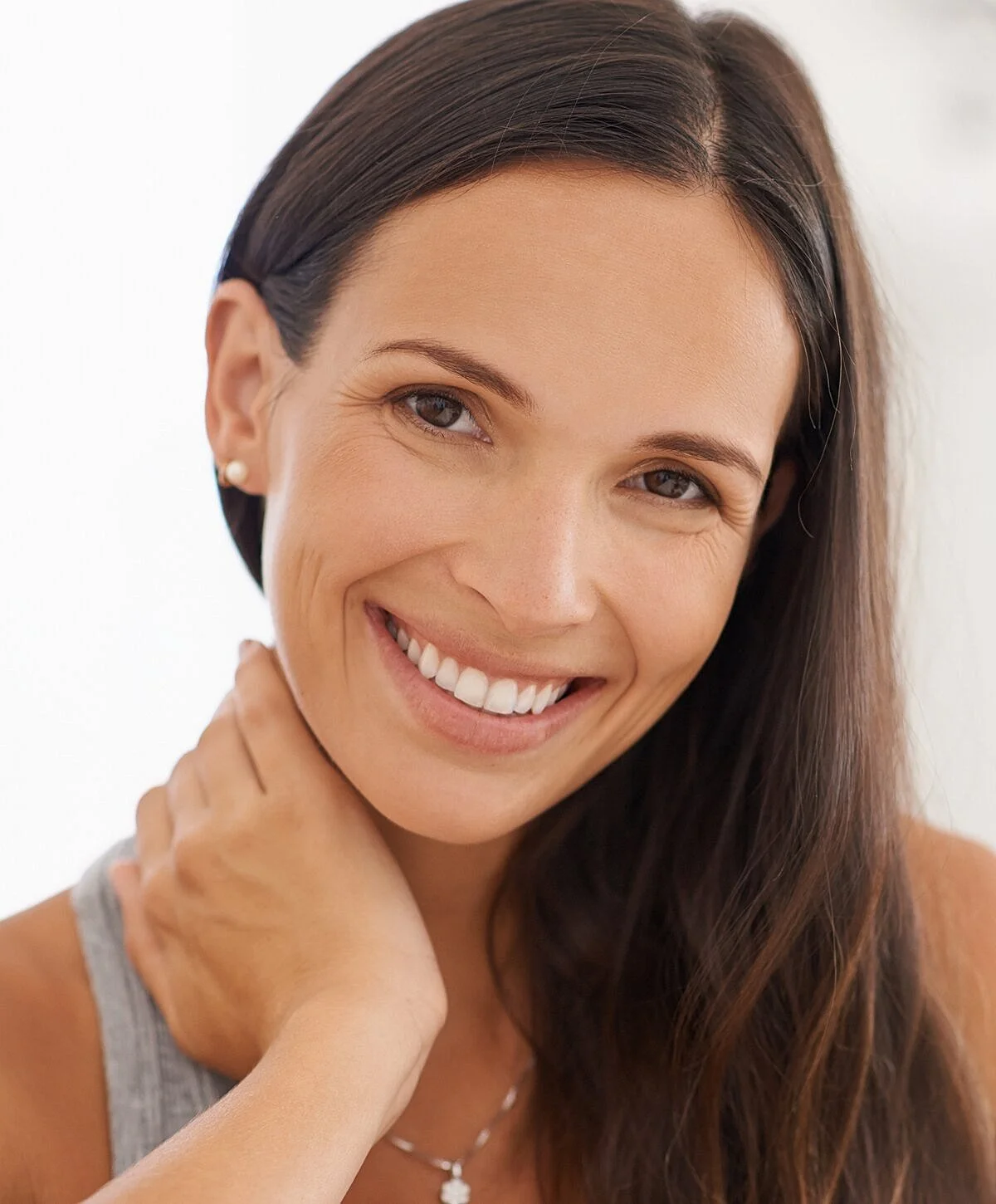 Close-up of a smiling woman with long brown hair, touching her neck, wearing pearl earrings and a necklace, against a plain white background.