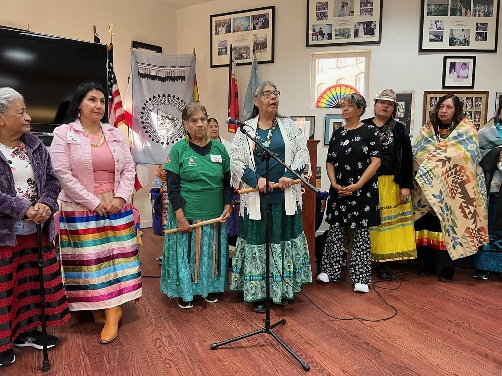 Group of women standing together at an indoor event, some dressed in traditional Native American attire, with flags and framed photos on the wall.