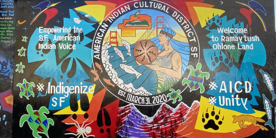 A colorful mural celebrating the American Indian Cultural District SF, featuring Indigenous symbols, a woman holding pottery, animals, and activist messages like 'Empowering the SF American Indian Voice,' '#IndigenizeSF,' and '#AICDUnity' with mountains and the Golden Gate Bridge in the background.