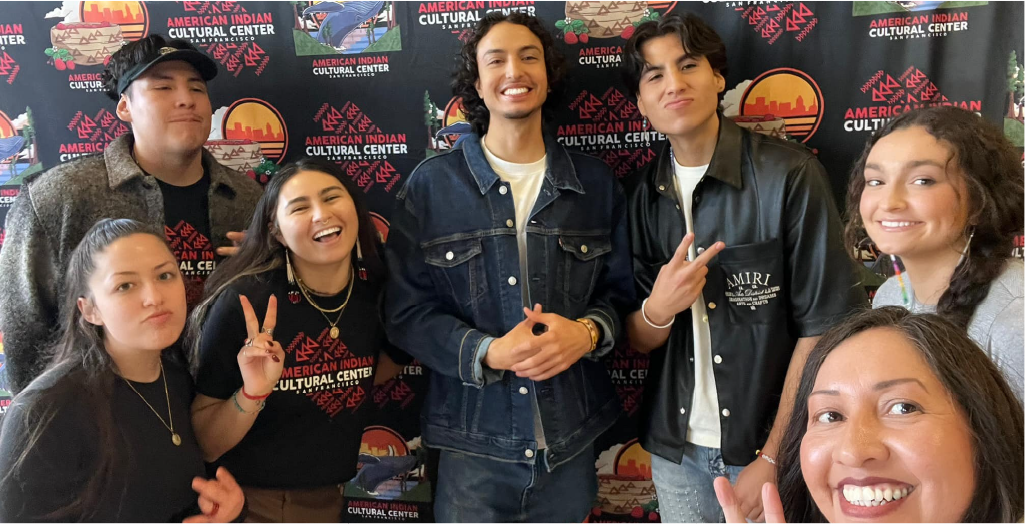 Group of seven young people smiling and posing in front of a backdrop for the American Indian Cultural Center, some making peace and victory signs and others smiling directly at the camera.