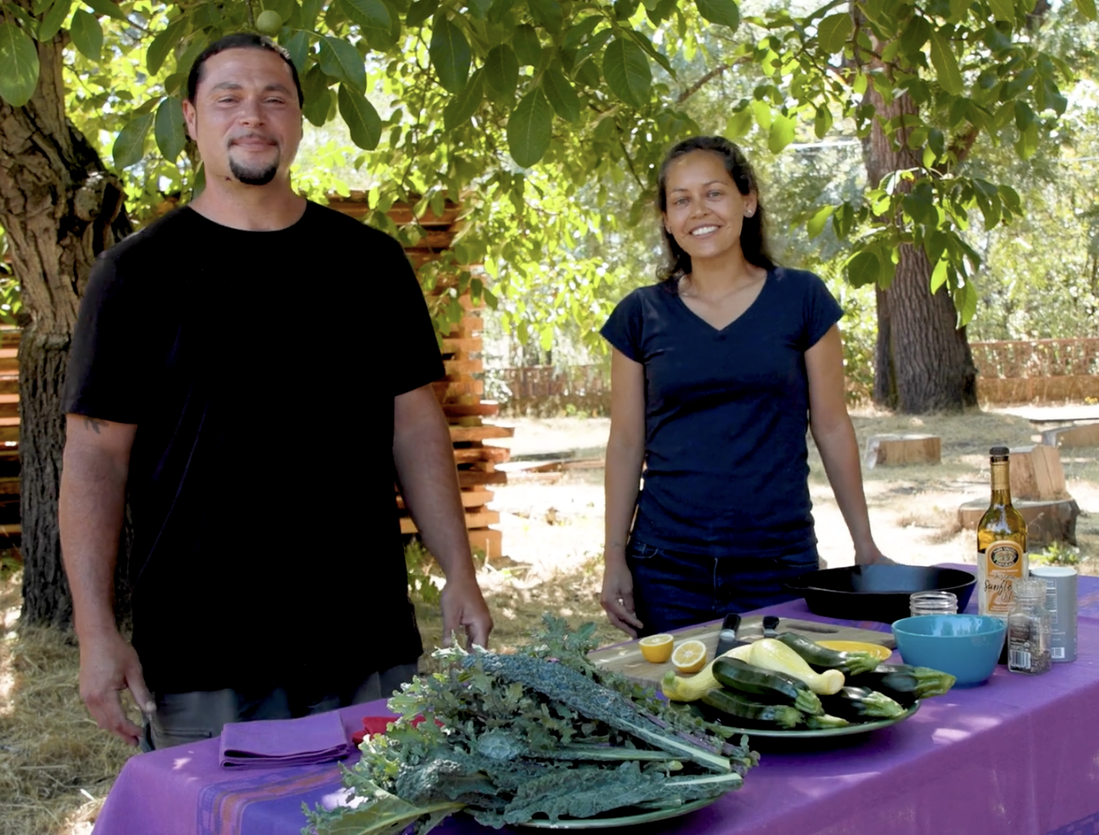 A man and woman standing outdoors behind a table with vegetables and cooking ingredients, smiling at the camera. The table has a purple tablecloth, with zucchini, yellow squash, and herbs, along with cooking utensils, a bottle of alcohol, and a blue bowl. The background features trees and a carved wooden fence.