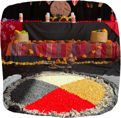 Decorative setup for a Diwali celebration with a large rangoli made of colored powders, a tiered altar with traditional sweets, candles, and decorative items, and festive ornaments and flowers in the background.