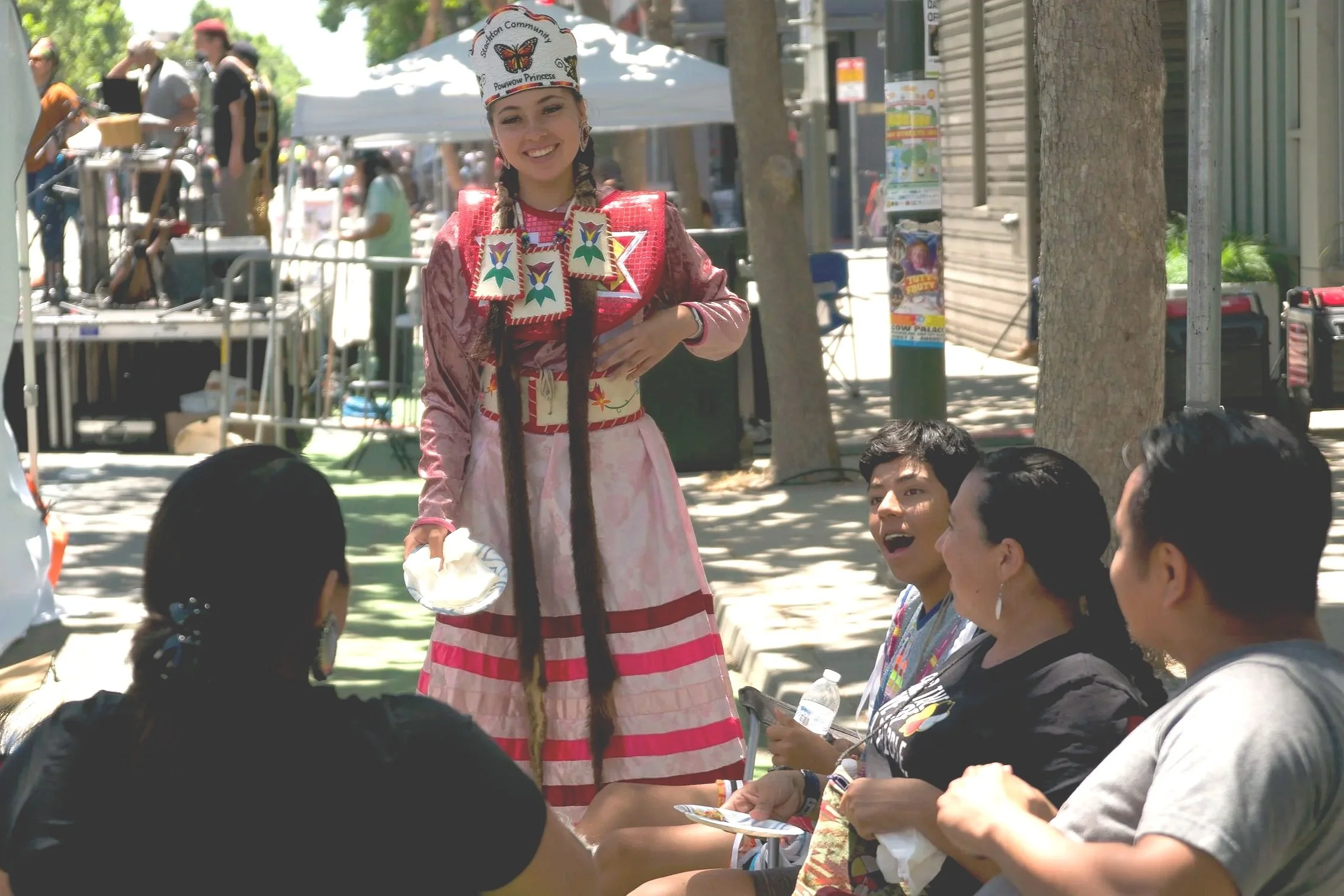 A young woman dressed in colorful Native American-inspired costume standing and smiling while talking to a group of four seated people outdoors during a community event.