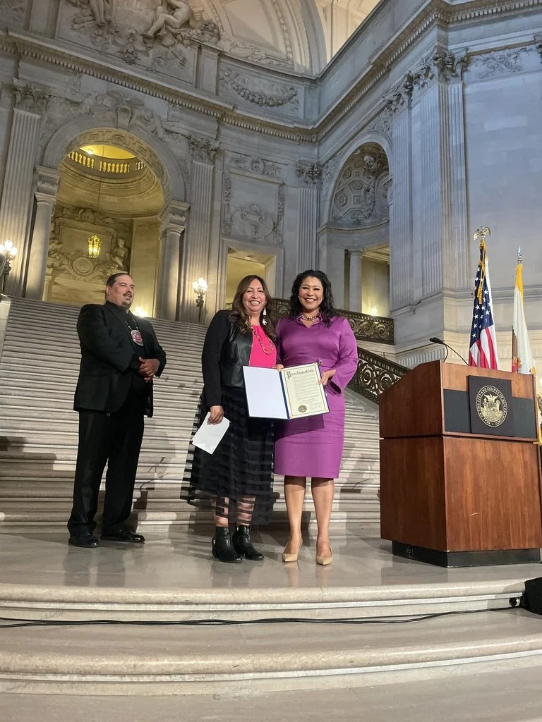 Two women standing on a grand staircase holding a certificate, smiling for the camera, with a man standing nearby in a formal setting inside a historic government building.