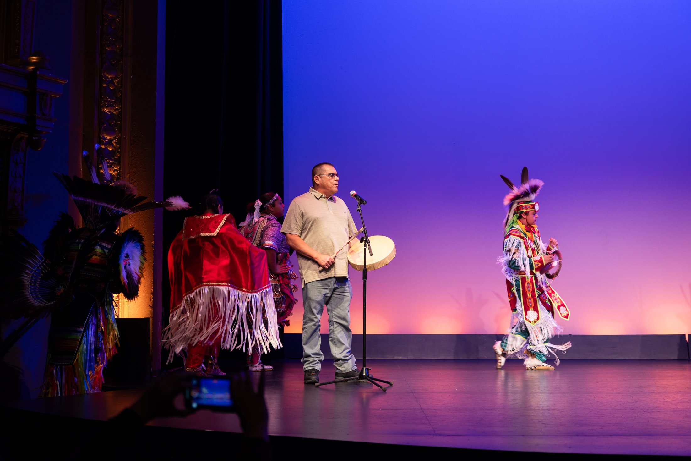 A stage performance with Native American dancers in traditional regalia, a man holding a drum and drumstick, and a colorful backdrop in purple and orange hues.