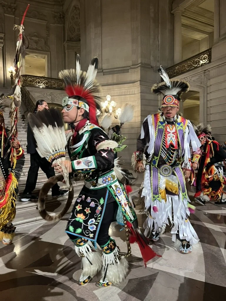 Indigenous dancers dressed in traditional, colorful regalia with feathered headdresses, performing inside an ornate building.
