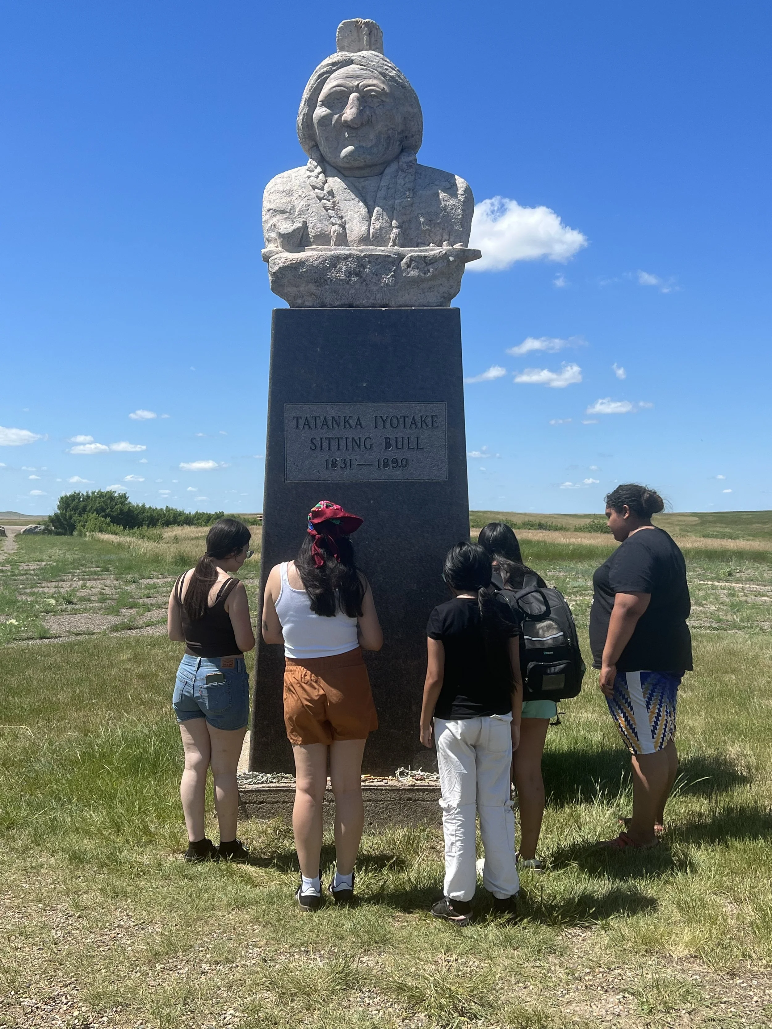 A group of five people gathered around a large statue of a sitting bull with a plaque that reads 'Tatanka Iyotake Sitting Bull 1831-1890' on a pedestal, outdoors on a bright, sunny day with a blue sky and some clouds.