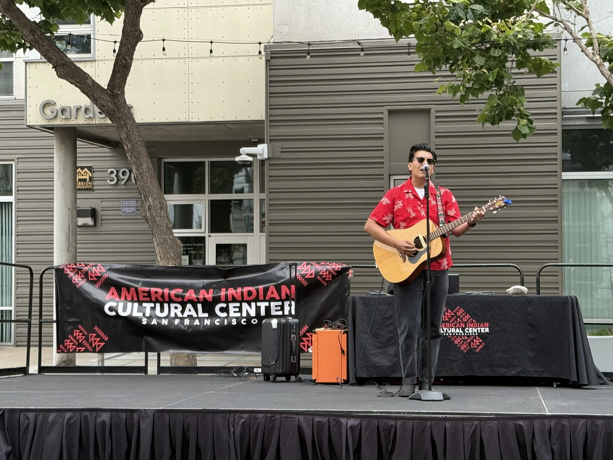 A performer at the American Indian Cultural Center's stage performing at Sunday Streets in San Francisco. There are trees and a modern building in the background, and a banner on the stage reads 'American Indian Cultural Center San Francisco'