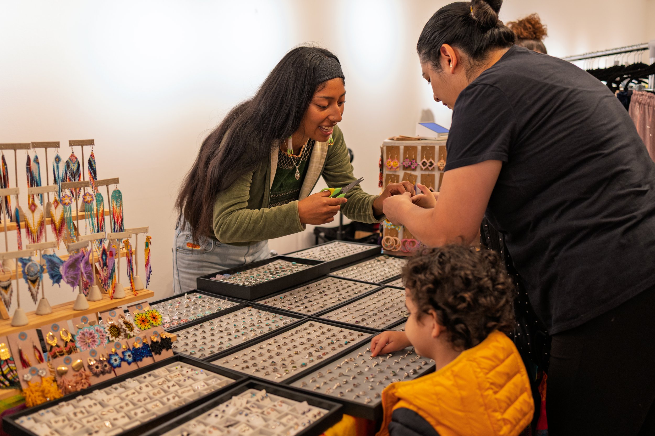Two women and a child shopping for jewelry at a vendor stall with various rings, earrings, and necklaces displayed.