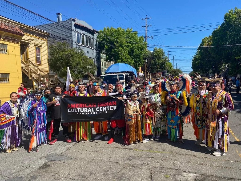 Group of people dressed in traditional Native American costumes holding a banner that reads 'American Indian Cultural Center San Francisco' during a parade on a sunny day.
