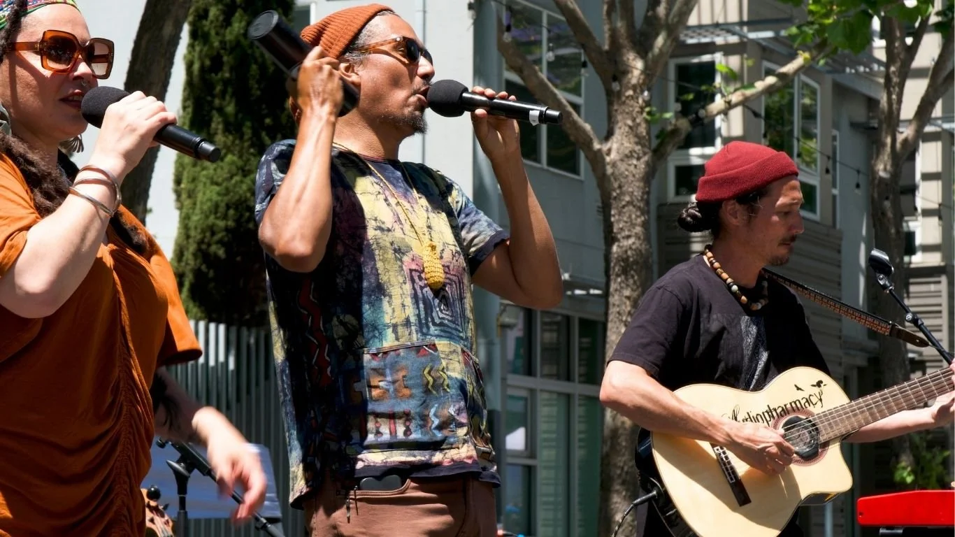 Three street performers singing and playing guitar outdoors, with trees and buildings in the background.