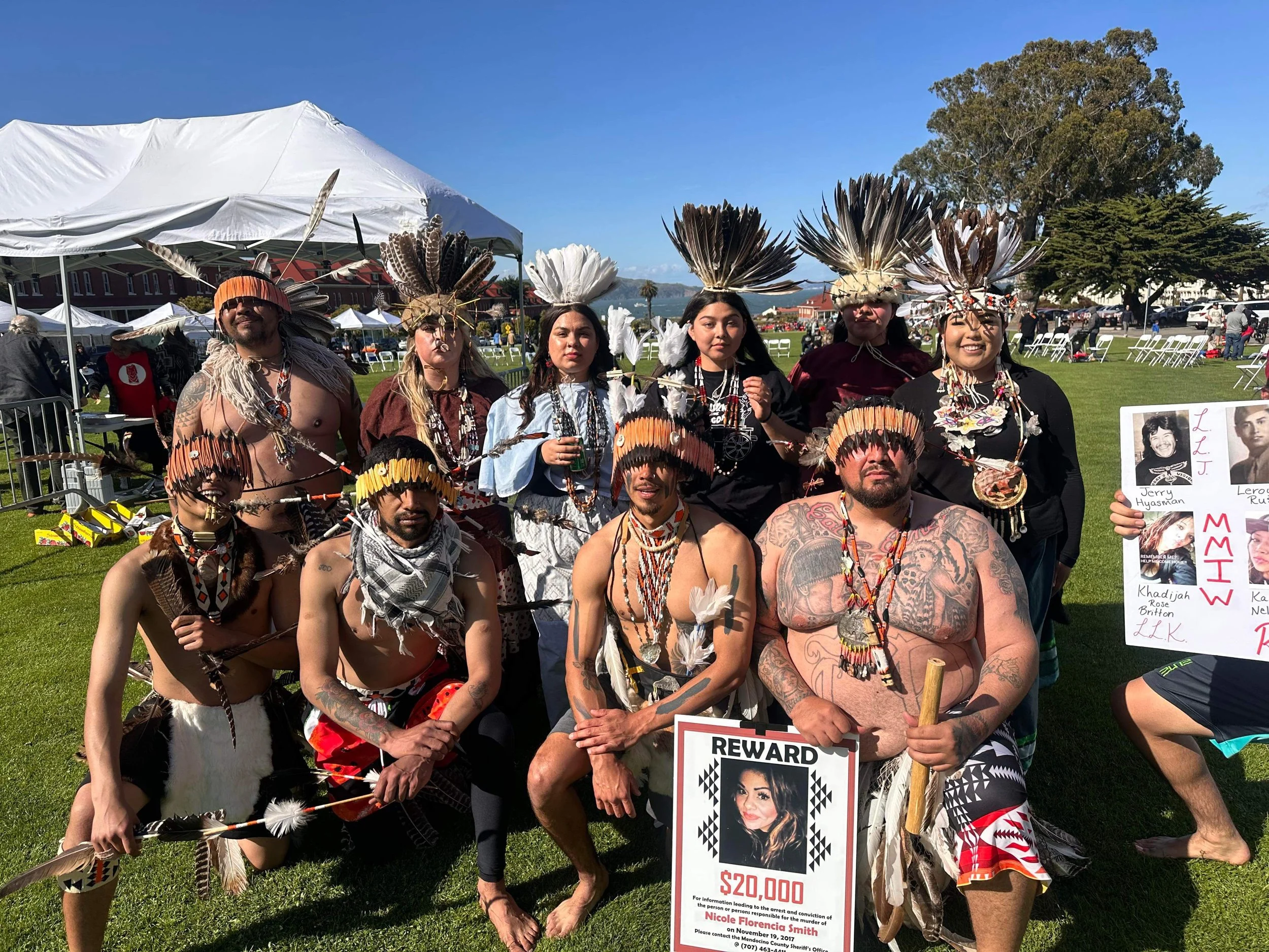 Group of indigenous people in traditional attire with feather headdresses, jewelry, and face paint, posing on a grassy field during a cultural event with a reward poster and informational board.