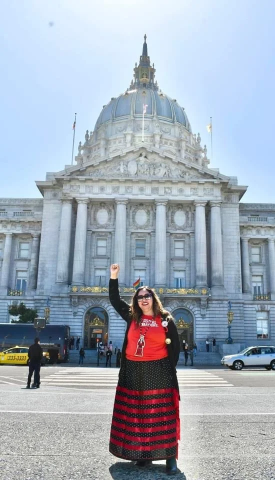 A woman with long curly hair wearing sunglasses, a red shirt, and a black and red striped skirt, standing in front of a historic white government building with a large dome, raising her fist and smiling on a sunny day.