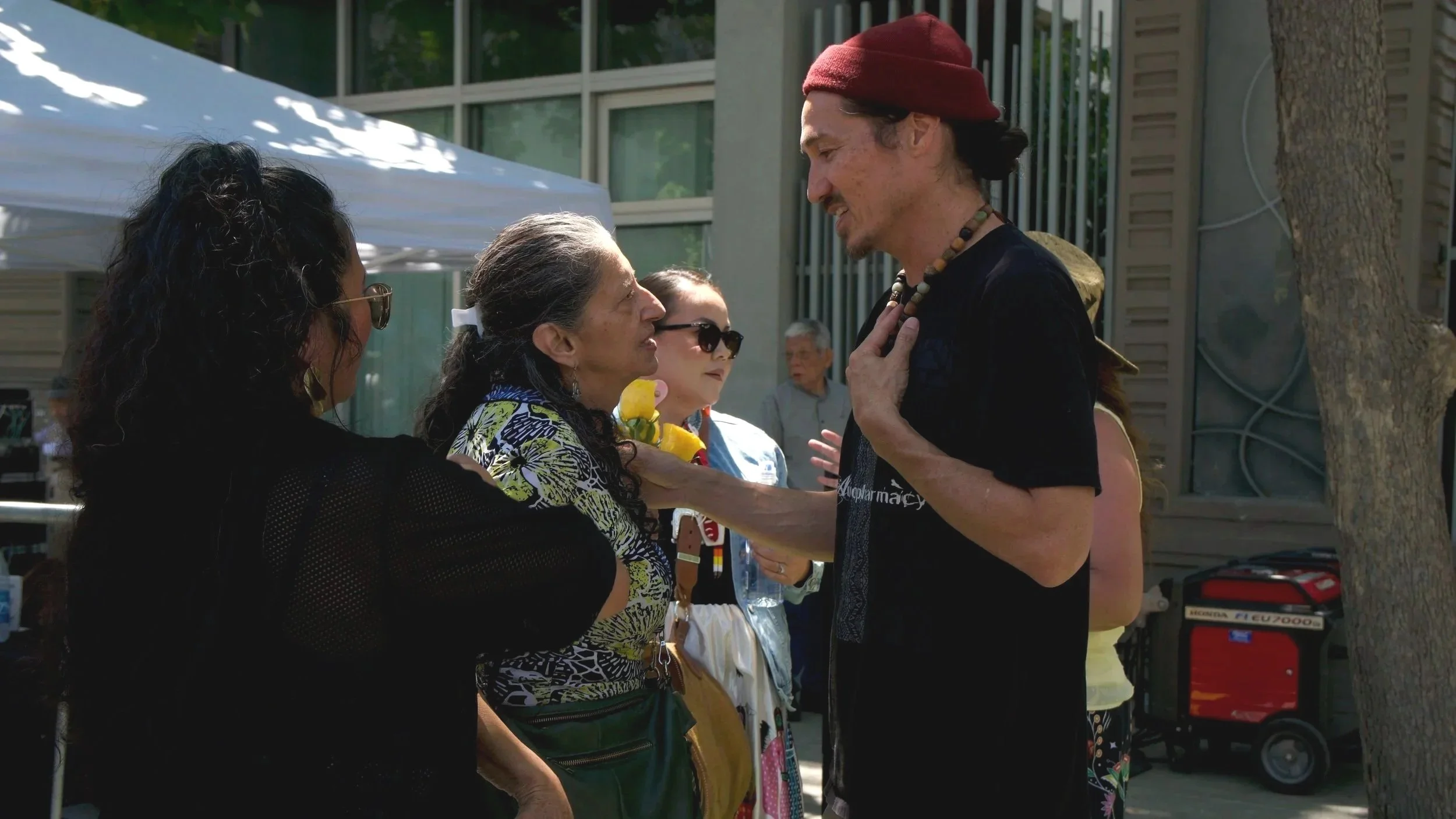 A man with long dark hair, wearing a red hat and black shirt, is talking to an older woman with gray hair and wearing a patterned blouse, at an outdoor event. Several people are in the background.