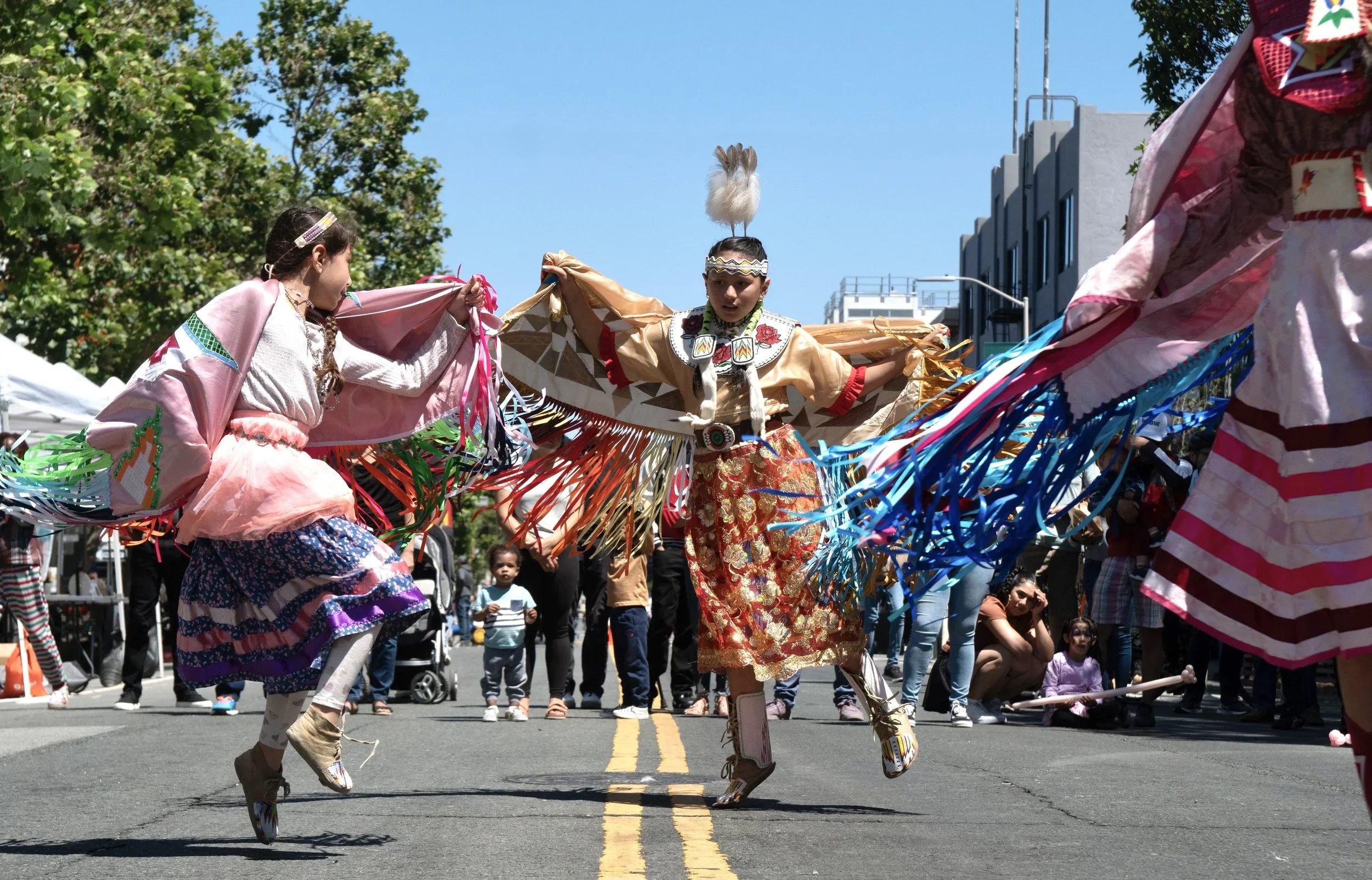 People participating in a cultural dance parade, dressed in traditional Native American attire, with some jumping and dancing on a city street, spectators watching from the sides.