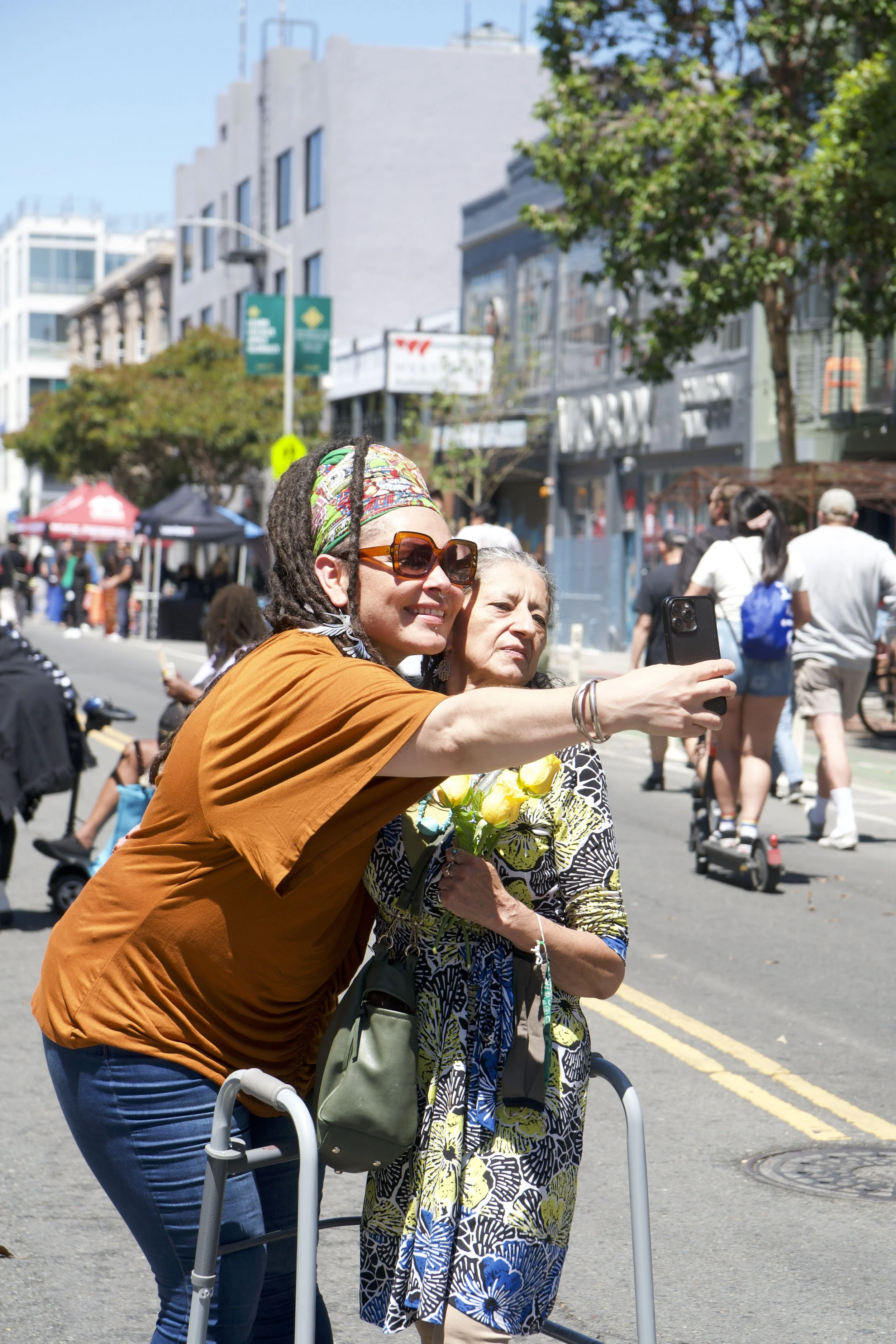 Two women, one with a walker, taking a selfie together on a busy city street during a sunny day.