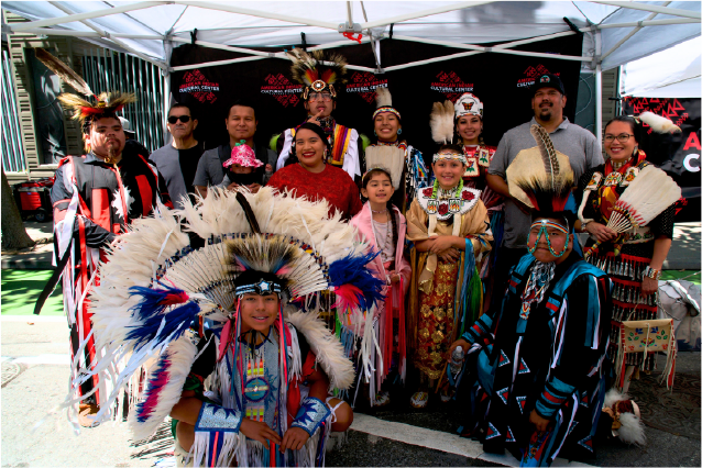 Group of people in traditional Native American attire, some wearing feathered headdresses, standing under a canopy at an outdoor event.