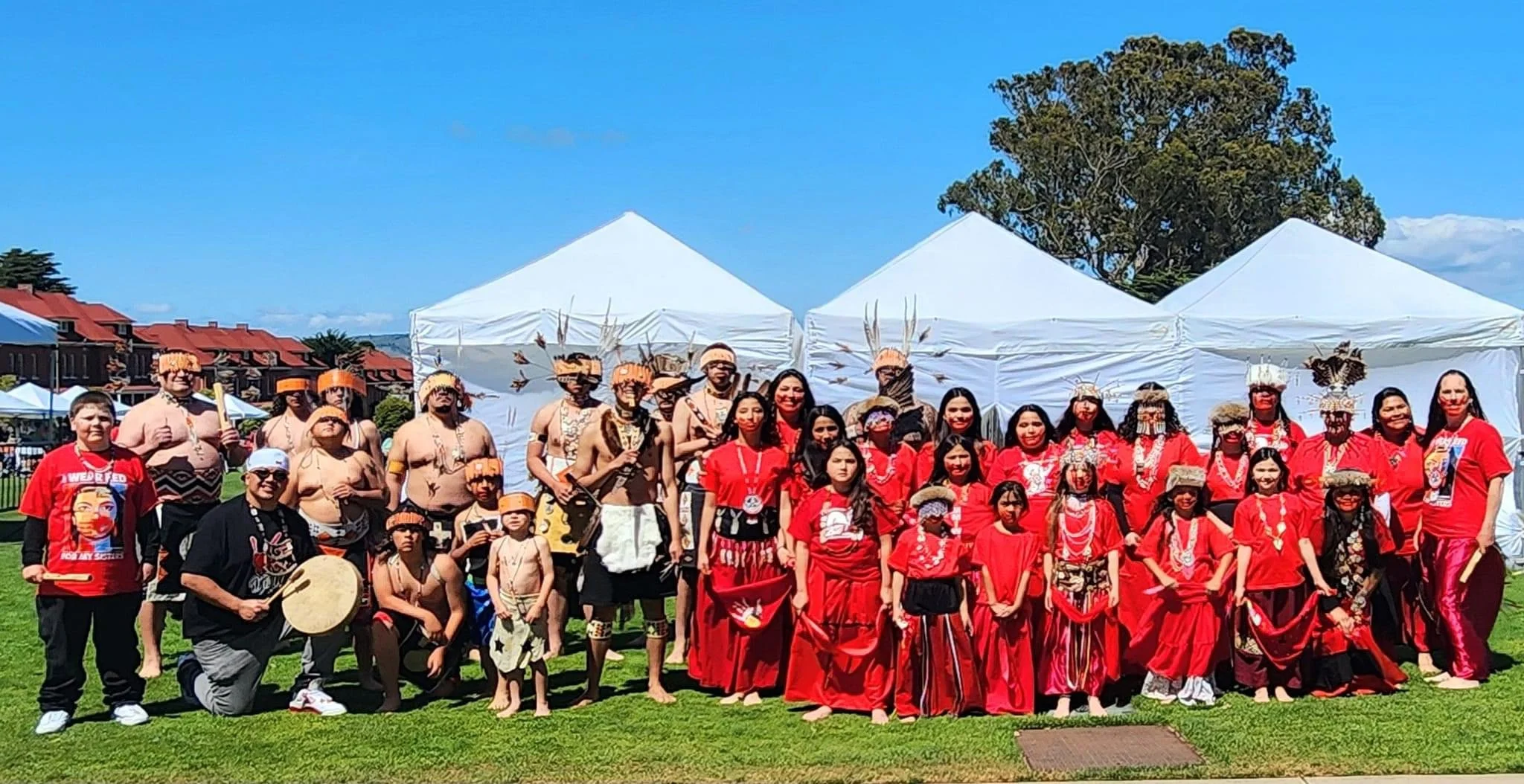 Group of Indigenous people and supporters in traditional attire and red clothing, posing outdoors under white tents on a sunny day.