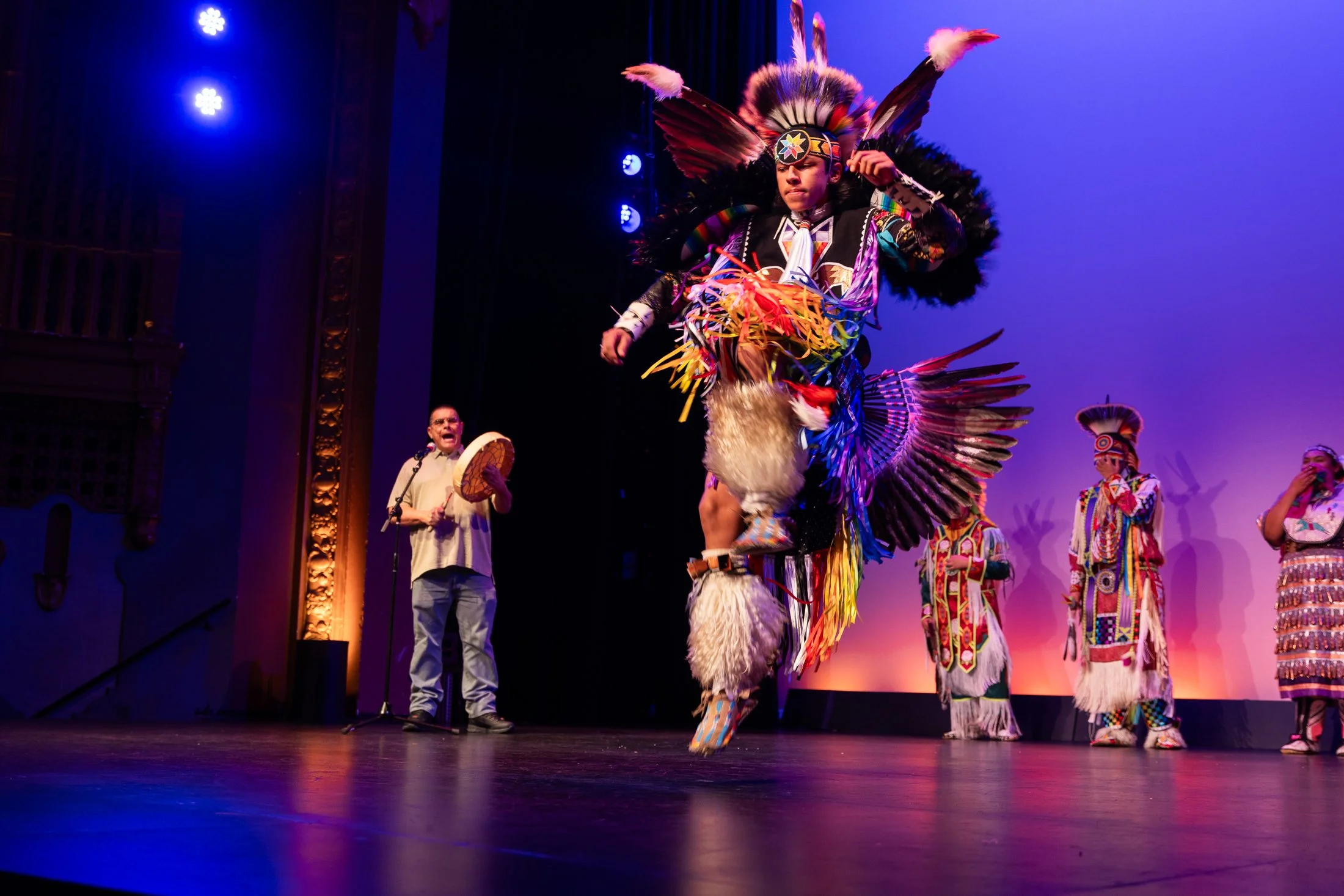 A performer dressed in a colorful indigenous costume, including feathers and traditional attire, performing a dance on stage. Other performers in similar costumes and a musician playing a drum are visible in the background.