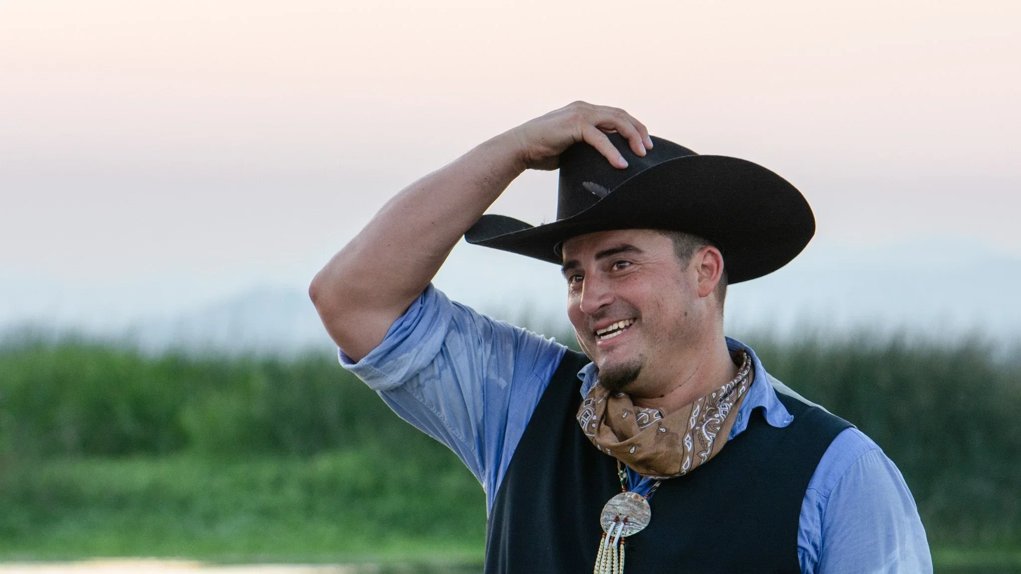 A man wearing a cowboy hat, blue shirt, and a bandana around his neck, smiling outdoors with a grassy field and trees in the background.