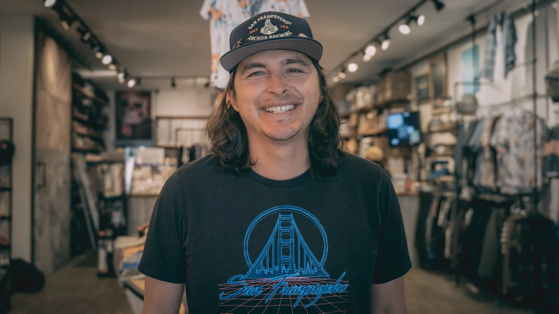 A man with long hair and a cap smiling in a store with shelves of merchandise in the background.