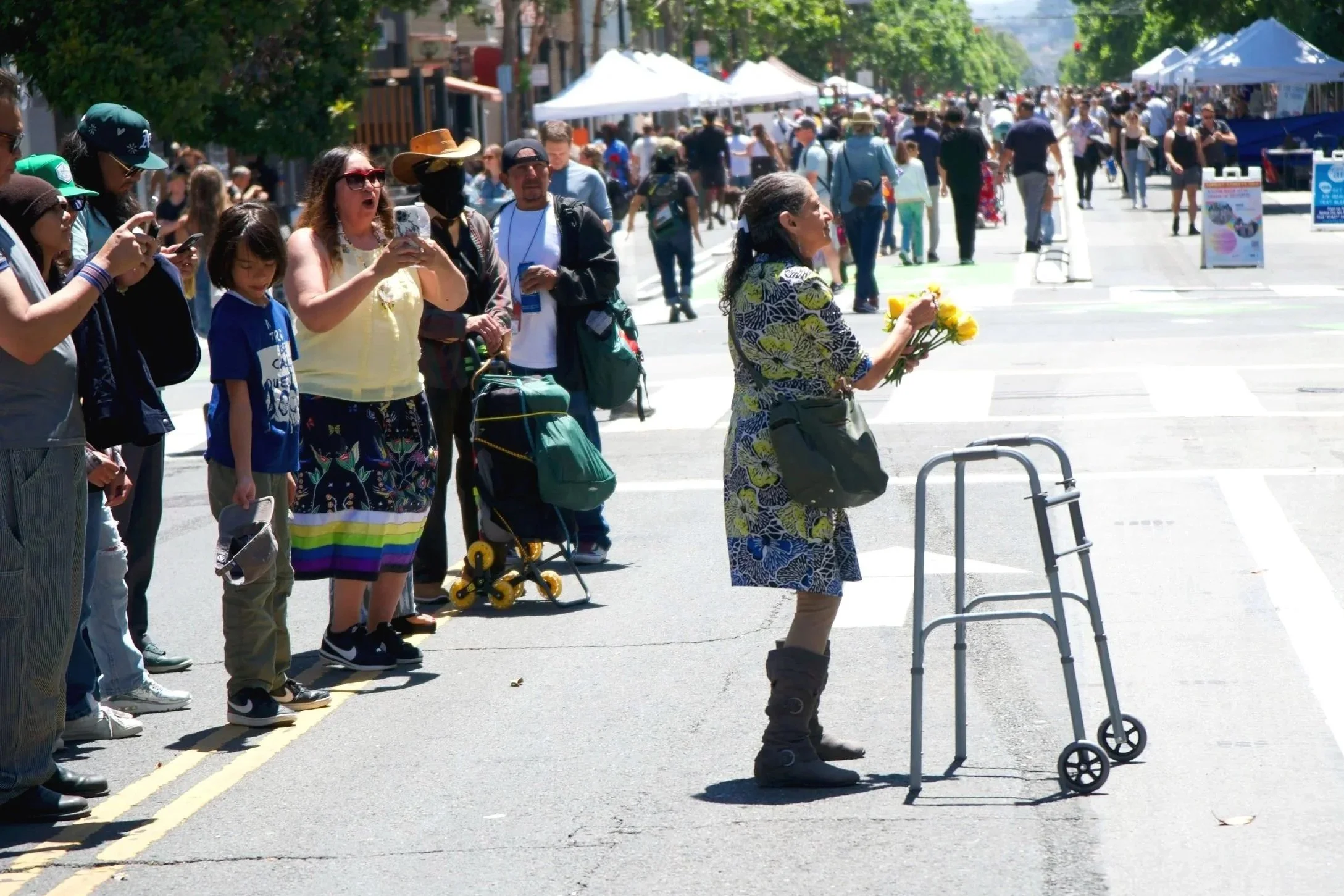 A woman with a bouquet of yellow flowers standing on the street, speaking to a crowd of people behind her during a street event or protest on a sunny day. The crowd is watching attentively, some recording with their phones, with tents and trees in the background.