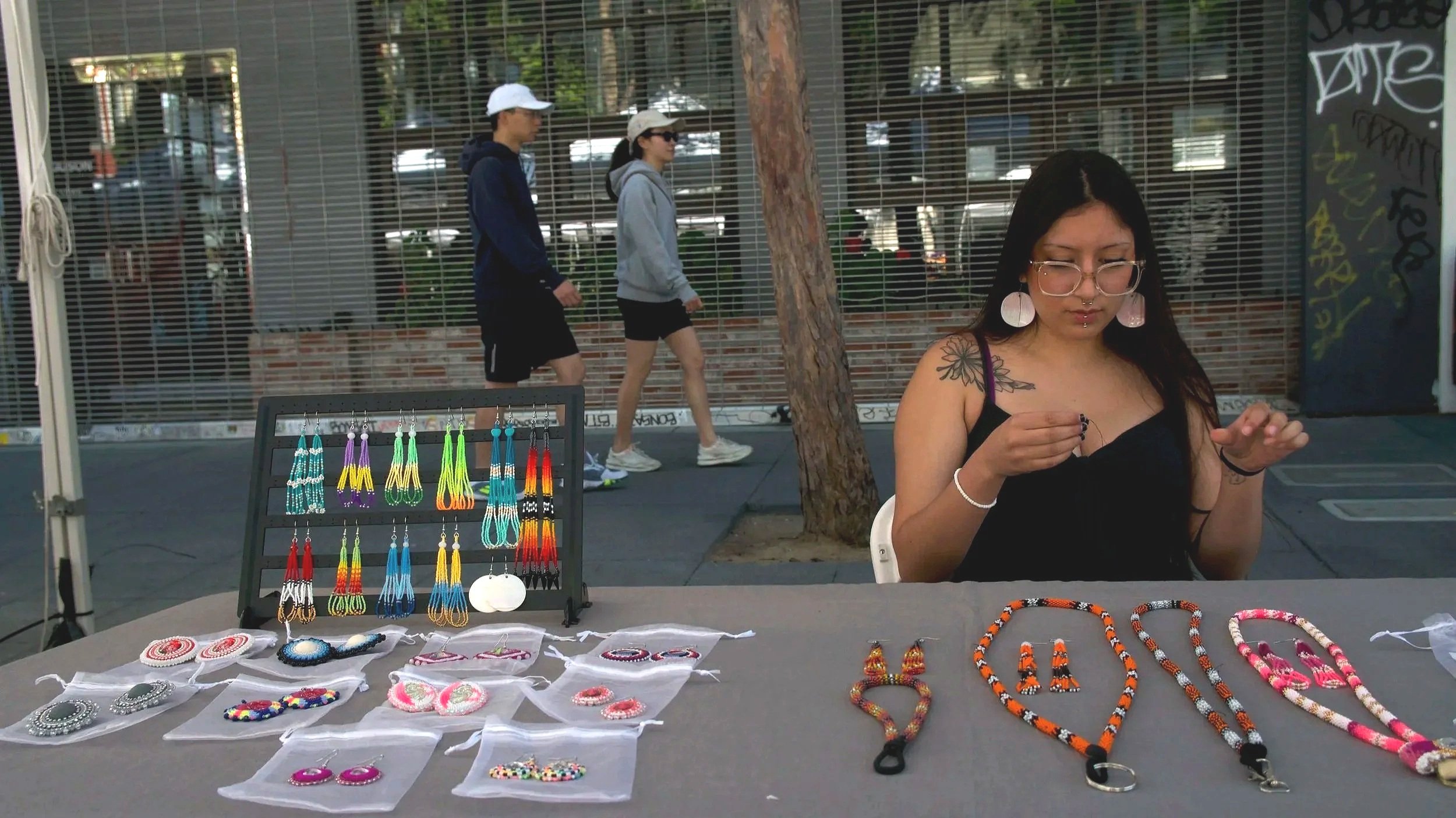 A woman sitting at a table displaying handmade jewelry including earrings, necklaces, and beaded accessories for sale at an outdoor market.