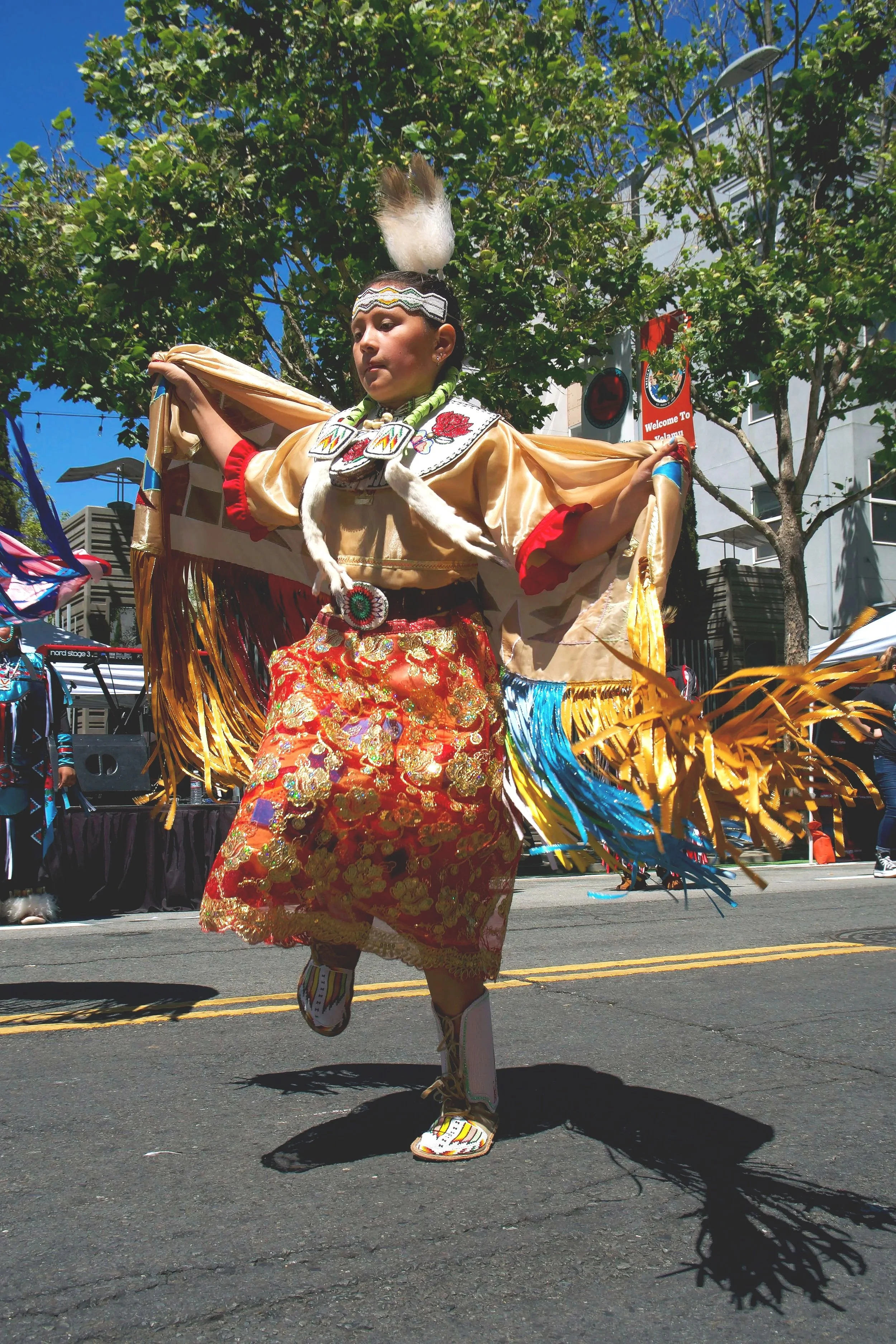 A Native American dancer performing in a parade, dressed in traditional clothing with detailed embroidery, fringes, and accessories, holding his arms out as he dances on the street under a blue sky.