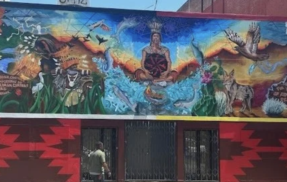 Colorful mural depicting indigenous woman seated cross-legged with a shield, surrounded by animals like wolves, deer, owls, and birds, with a vibrant landscape and sky background.