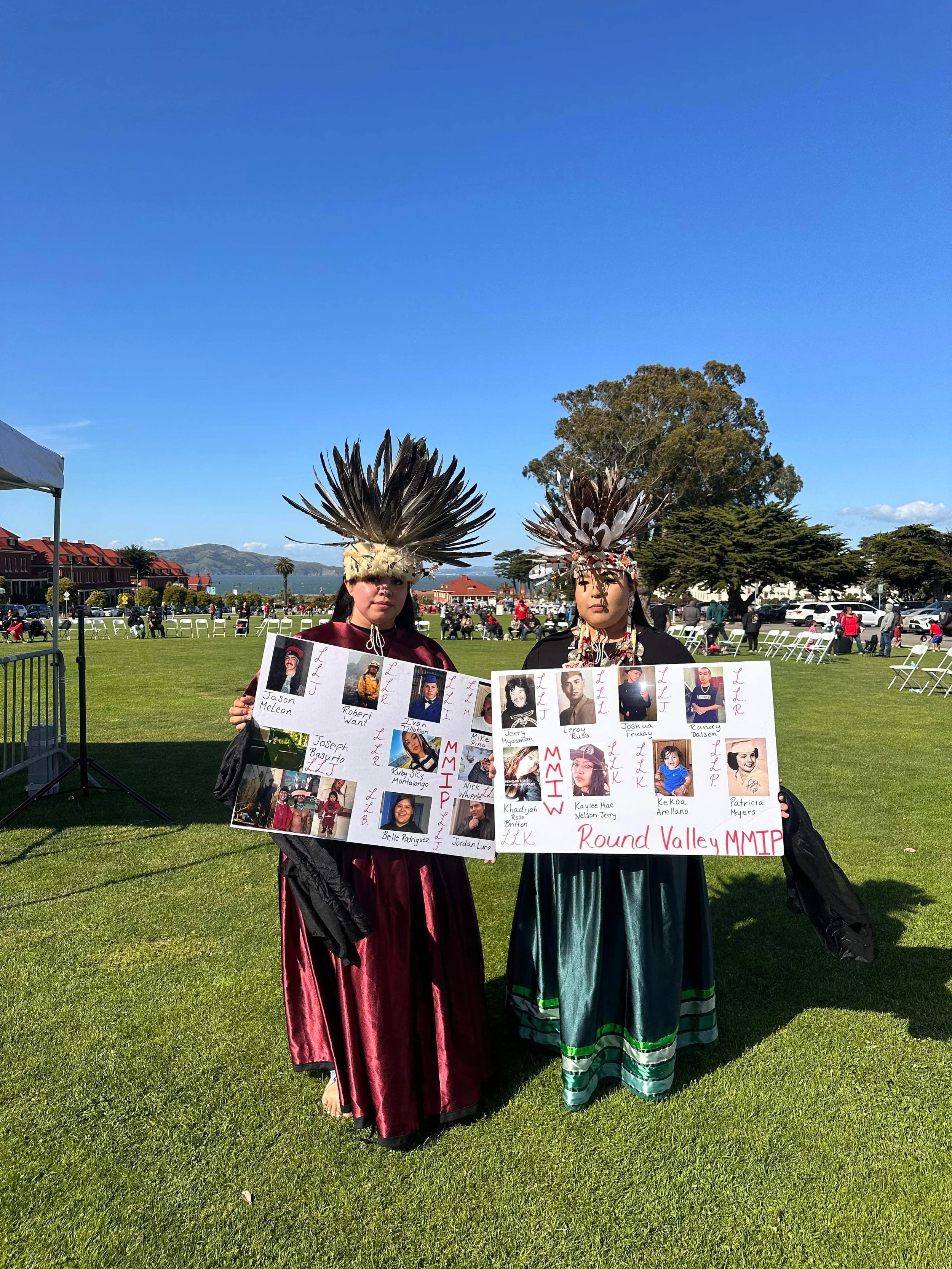 Two women dressed in traditional Indigenous attire holding a poster with photos and names of individuals, standing on a grassy area during a public event.