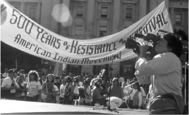 A protest with a large banner reading '50 Years of Resistance and Festival' and a crowd of people, some holding signs, gathered in front of a historic building. A person is filming the event with a video camera.