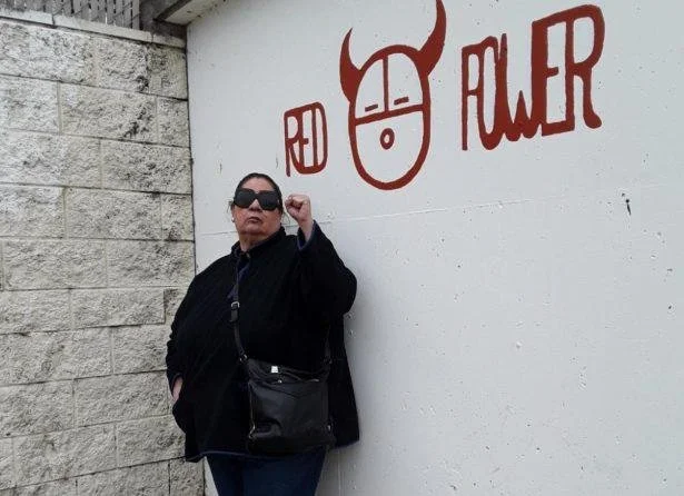 Person wearing sunglasses and black outfit standing against a wall with a red mural of a bull's head and the words "Red Fluer."