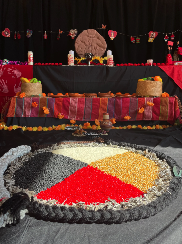 Colorful altar decorated for Day of the Dead, featuring a large round sugar or rice colored in black, red, yellow, and white, with offerings of fruits, candles, and marigold flowers, against a black backdrop with hanging paper hearts and butterflies.