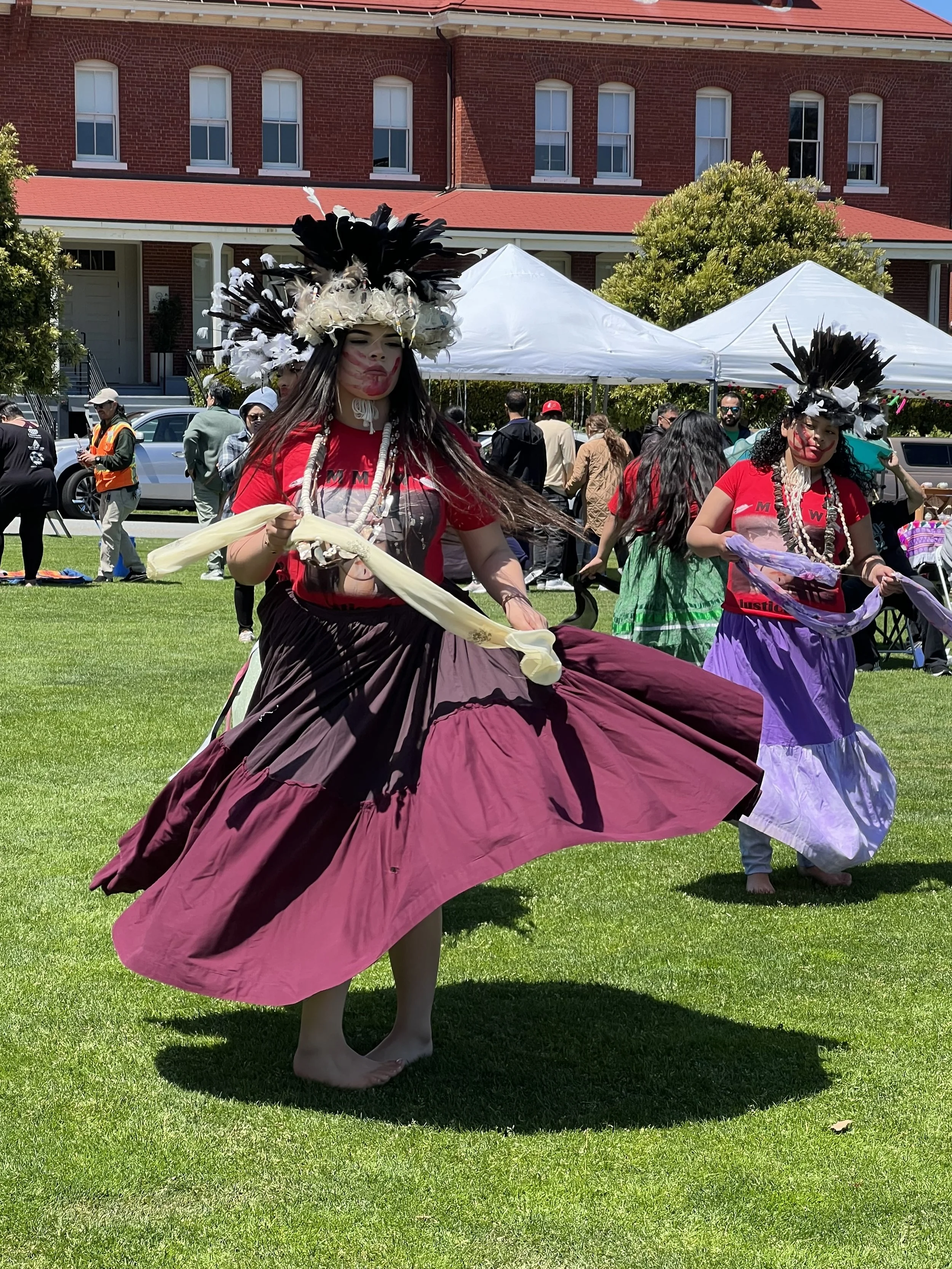Two women dressed in traditional Hawaiian attire dancing outdoors during a cultural event, wearing flowy skirts, feathered headdresses, and face paint, with tents and onlookers in the background.