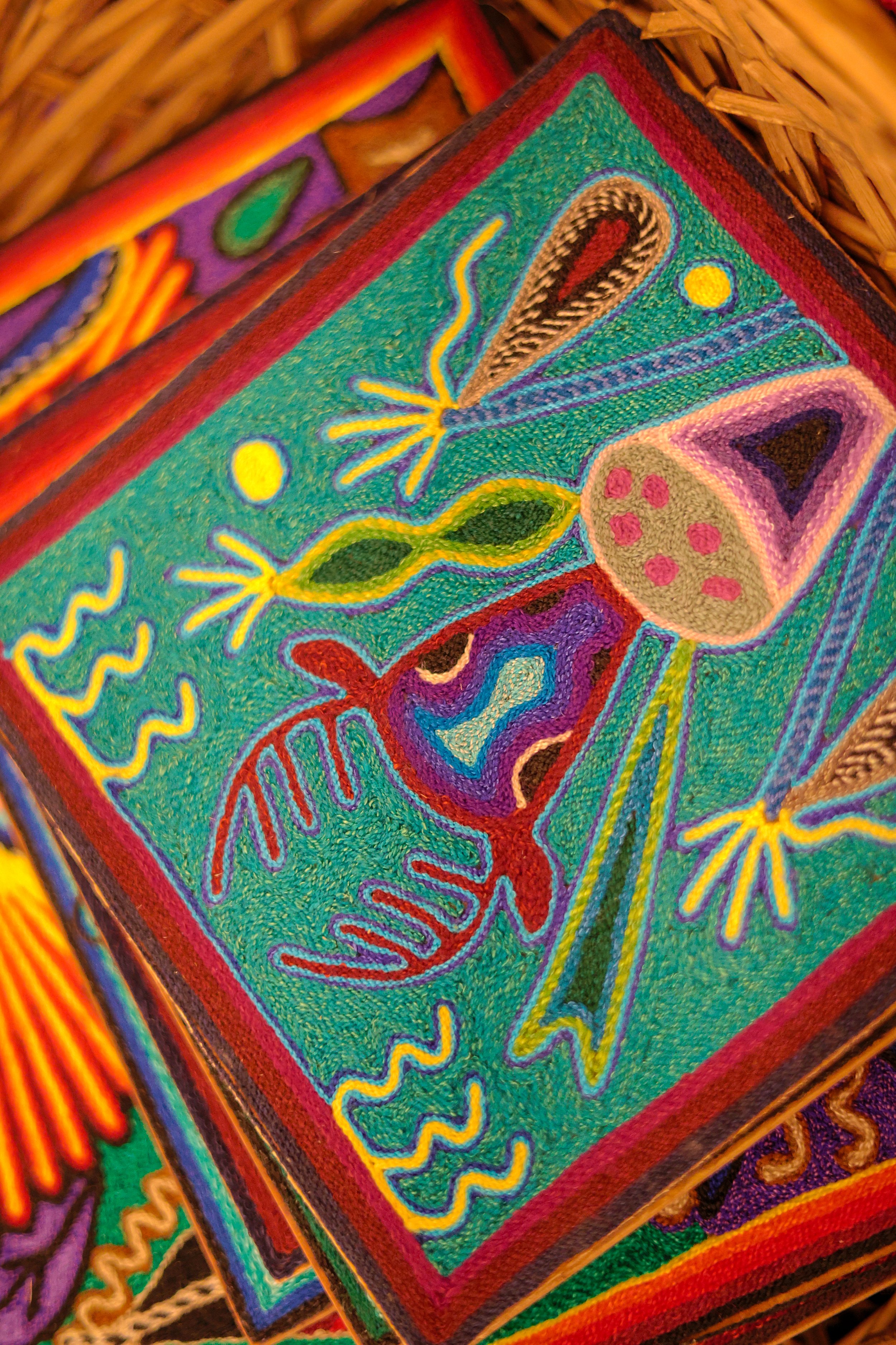 Close-up of colorful embroidered textile artwork featuring abstract shapes and patterns, including what appears to be a stylized ladybug and other organic forms, within a wooden basket.
