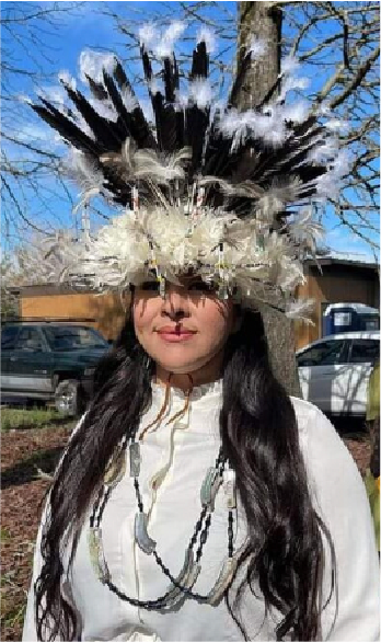 A woman wearing a large feathered headdress with black and white feathers, standing outdoors with trees and cars in the background.