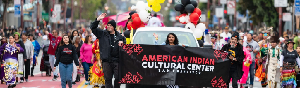 A parade with people marching down a city street, carrying a banner for the American Indian Cultural Center in San Francisco, with festive balloons in the background.