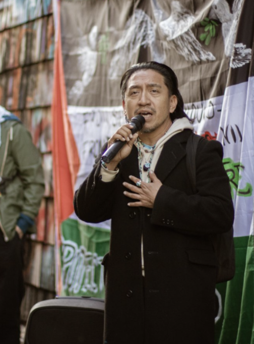 A man with long dark hair and a goatee singing into a microphone, wearing a black coat and gesturing with his right hand on his chest, standing outdoors in front of a banner and a group of people.