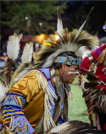 A Native American man dressed in traditional attire with feathered headdress, face paint, and beaded jewelry, participating in a cultural event or powwow.